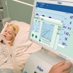 Ein großer Monitor in einem Krankenzimmer. Im Hintergrund sieht man eine Patientin im Bett liegen.